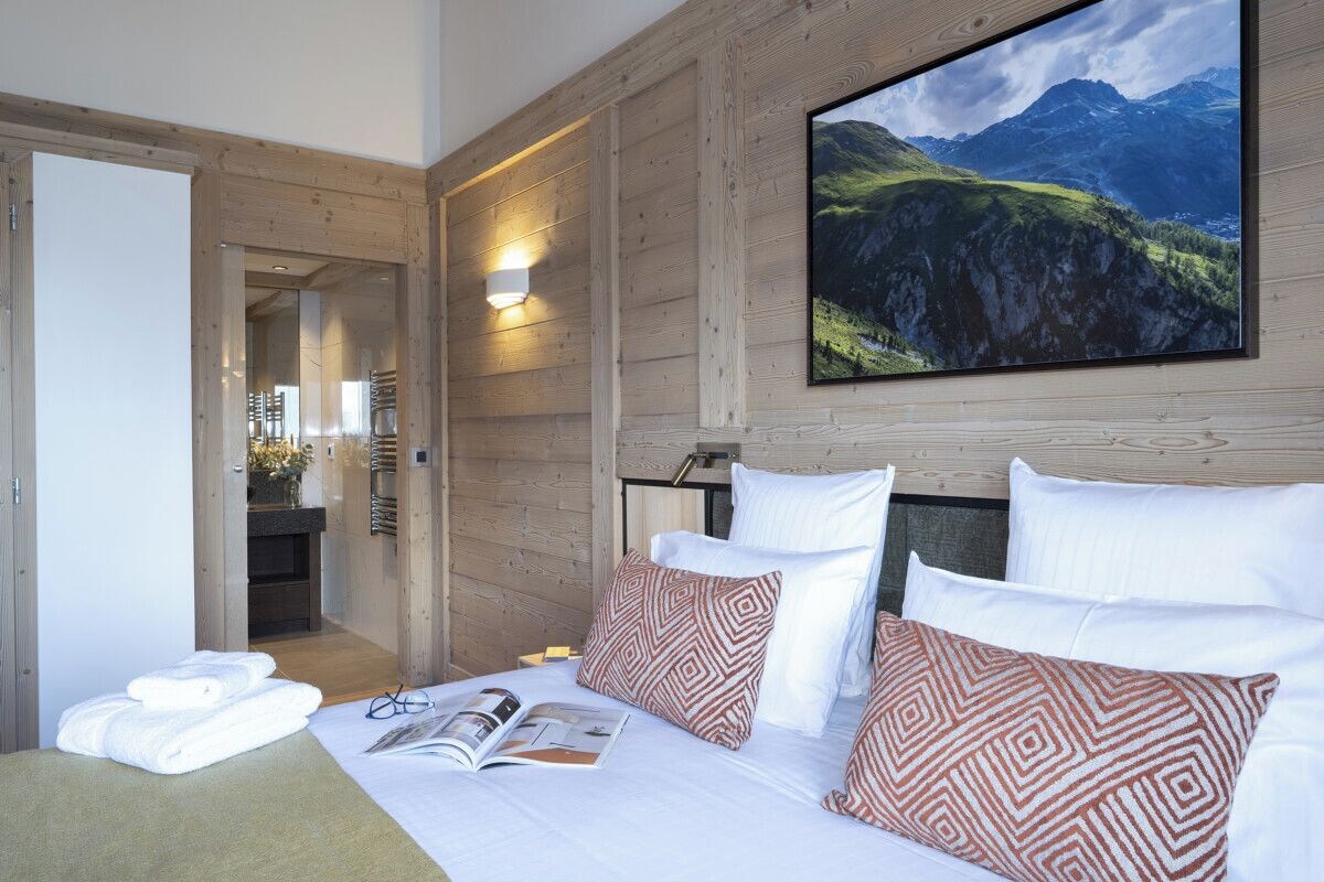 A hotel room with a bed, pillows, a magazine, and a large mountain view painting.
