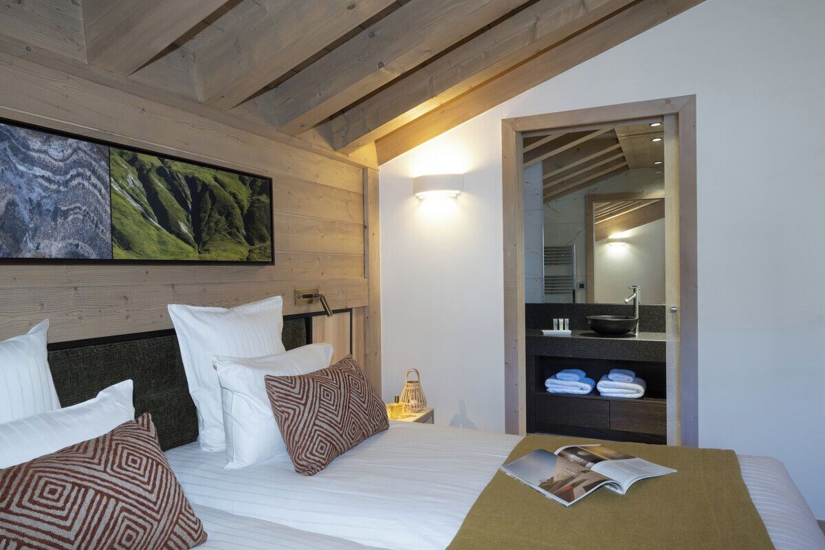 A bedroom with a bed, pillows, a nightstand, a wall-mounted light, a mirror, and a shelf with towels.