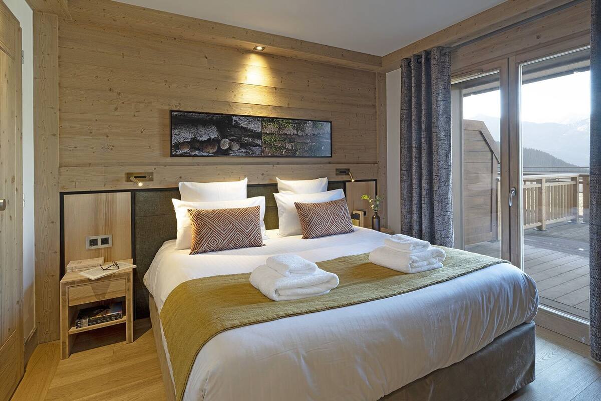 A hotel room with a large bed, a painting above the bed, a small bedside table, and a view of the outdoors through a sliding door.
