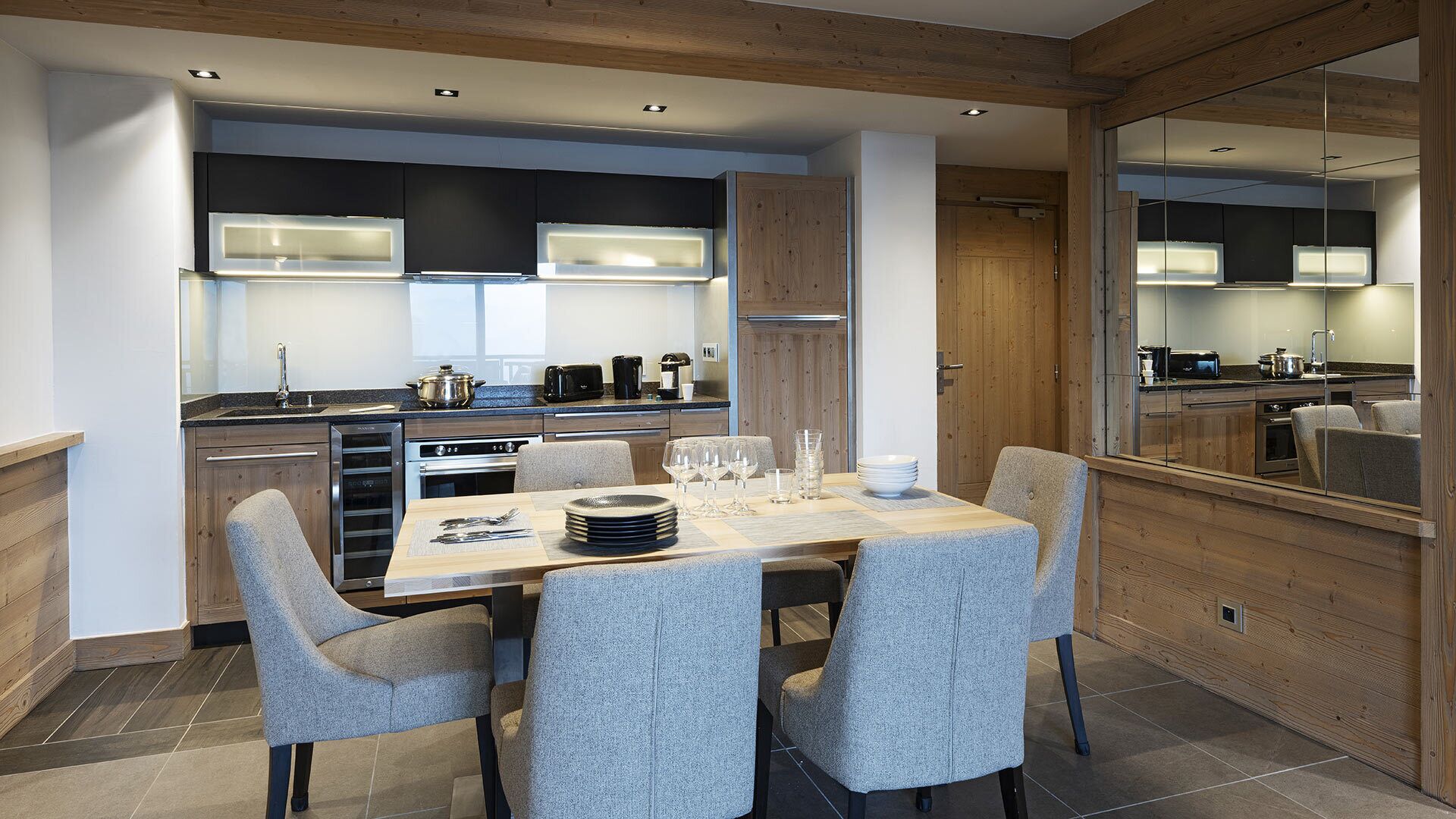 A modern kitchen with built-in appliances, a dining area with a table set for four, and a large mirror reflecting the kitchen.