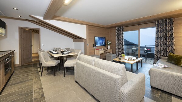 A modern living room with a sofa, dining table, and chairs. There is a TV mounted on the wall, and a balcony with a view. - MGM Hôtels & Résidences – Résidence Alpen Lodge (Montvalezan)