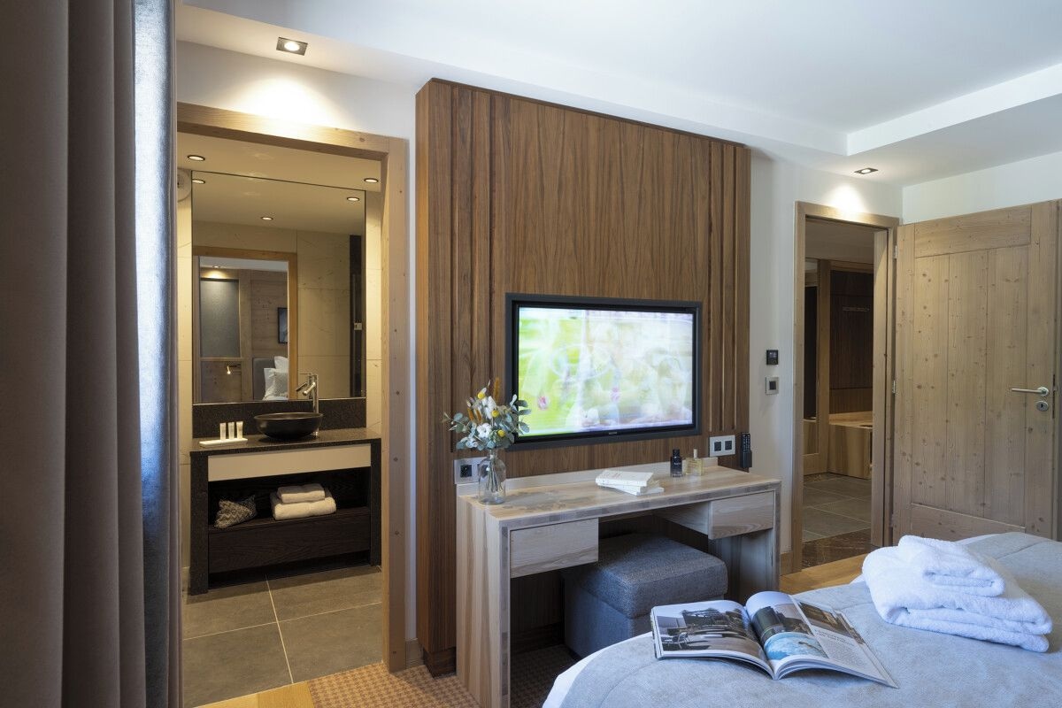 A modern hotel room with a wooden panel wall, a large flat-screen TV, a desk with a chair, a bed with a magazine and towels, and a bathroom visible through an open door.