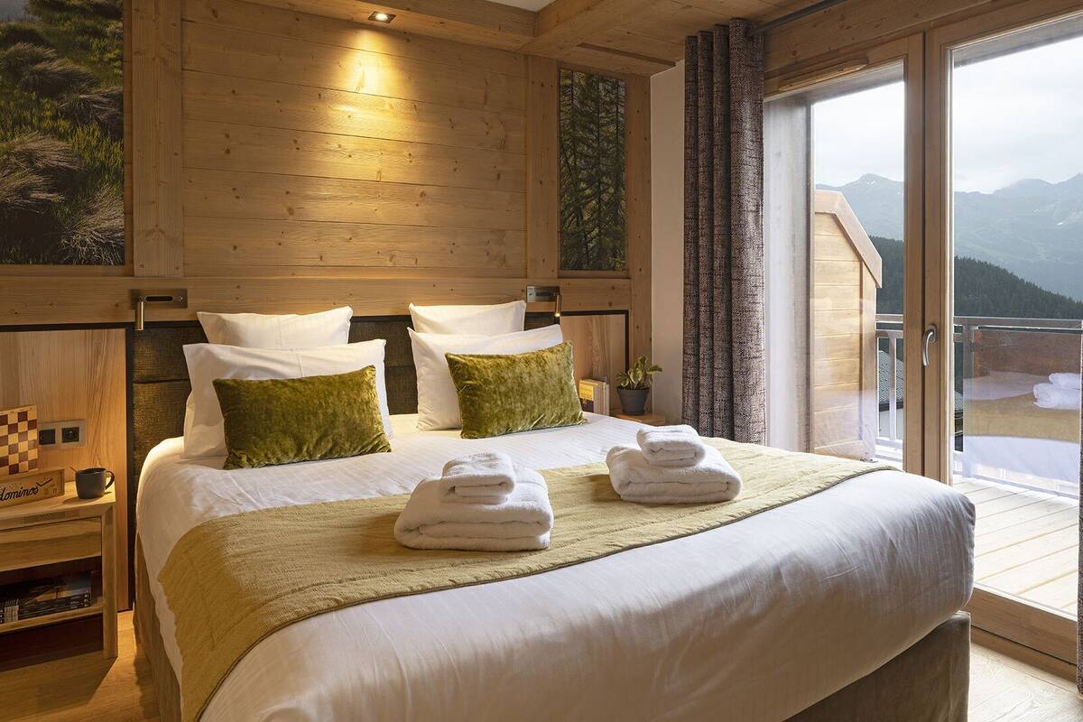 A wooden bedroom with a large bed, two pillows, a bedside table with a chessboard, and a view of mountains through a large window.
