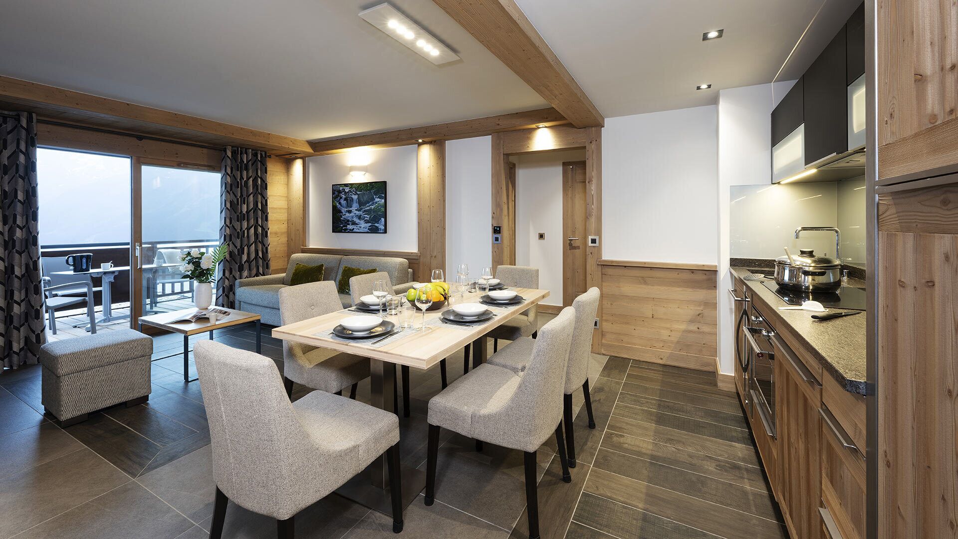 A modern dining area with a wooden table set for four, surrounded by cushioned chairs. A large window provides natural light, and a painting is visible on the wall.