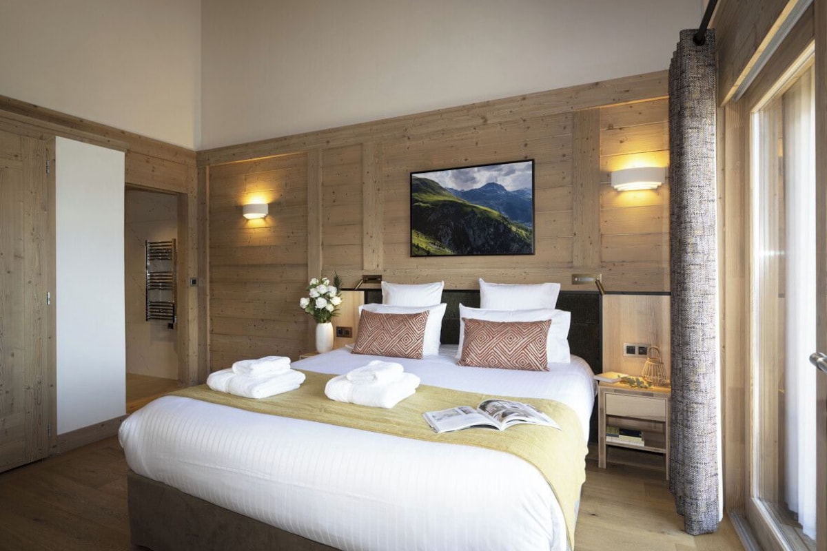 A hotel room with a large bed, wooden panel walls, a mountain landscape painting, and a window with curtains.