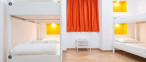 Une pièce avec des lits superposés, une petite table blanche, des rideaux orange et deux fenêtres éclairées d’une lumière jaune.