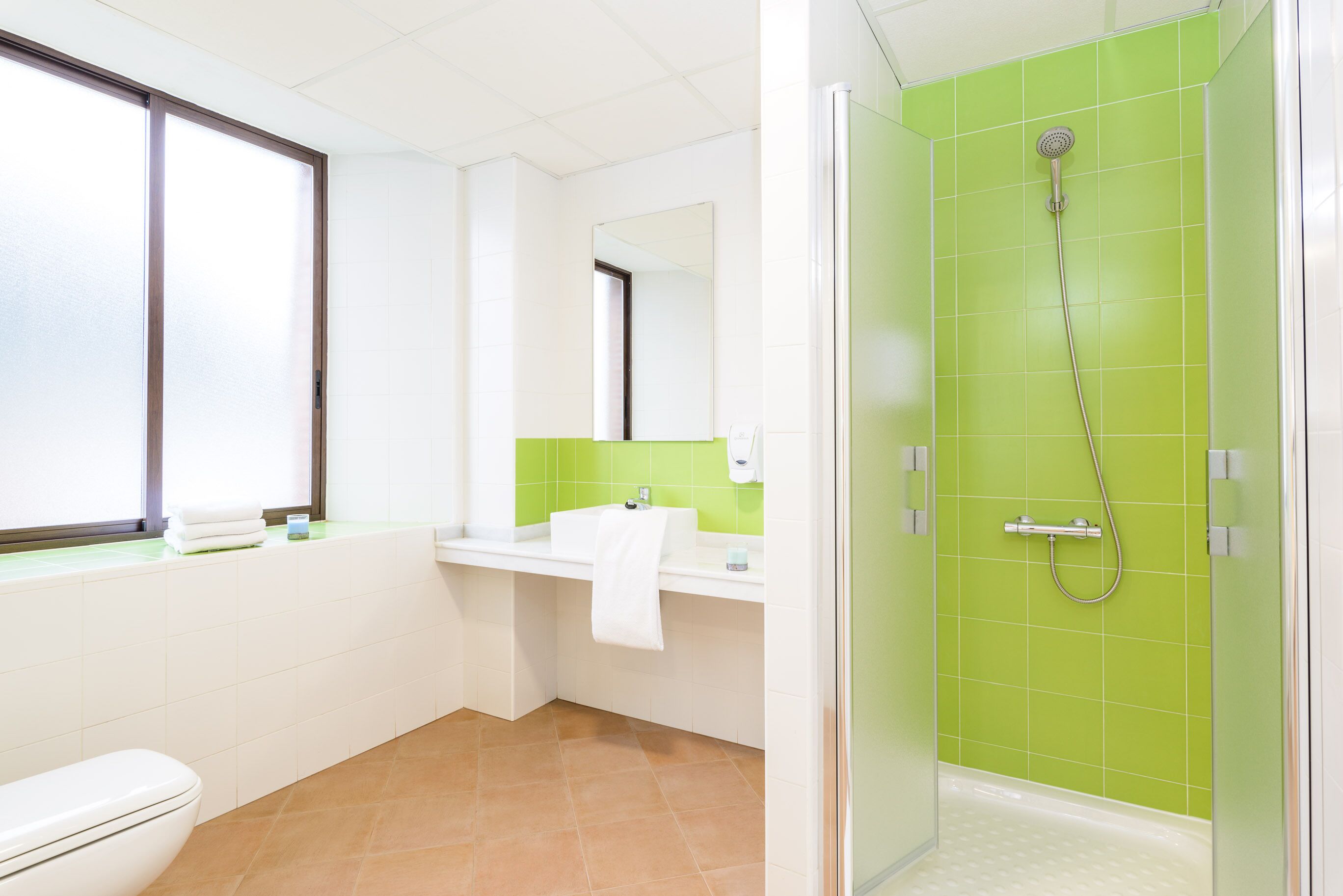Baño con inodoro, lavamanos, espejo y ducha con azulejos verdes.