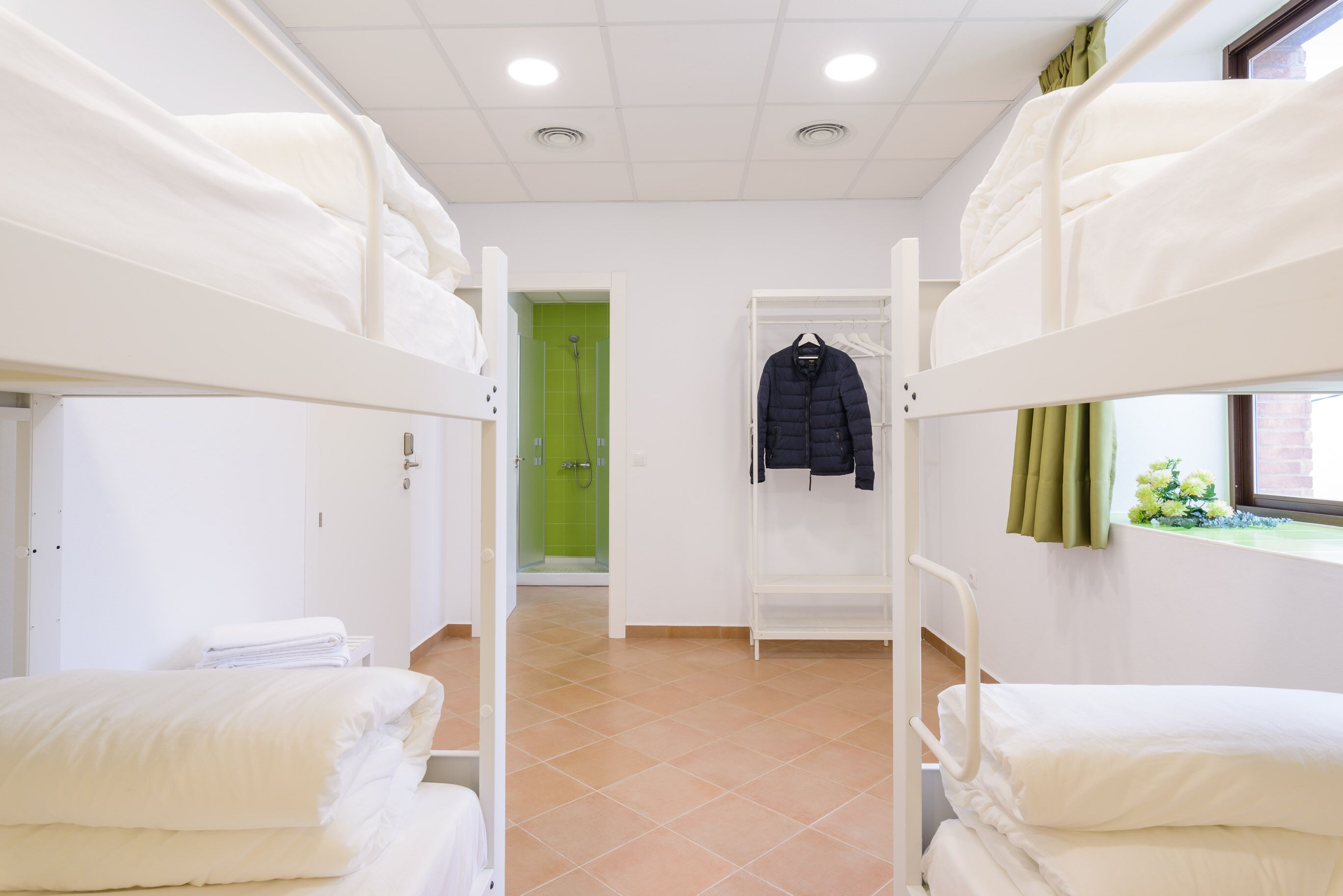 Une chambre avec des lits superposés, des draps blancs, une porte verte et une fenêtre avec un rideau.