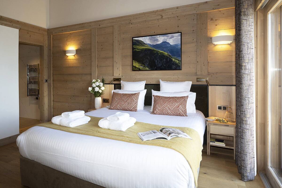Ein Hotelzimmer mit einer holzverkleideten Wand, einem Bett mit weißen Leinen, gemusterten Kissen, einer Vase mit Blumen und einem Gemälde einer Berglandschaft.