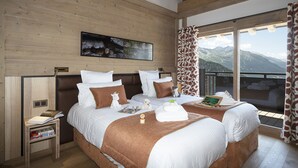 A double bed with white and brown bedding, a nightstand with books, a painting on the wall, and a view of mountains through the window. - MGM Hôtels & Résidences – Hôtel Alpen Lodge (Montvalezan)