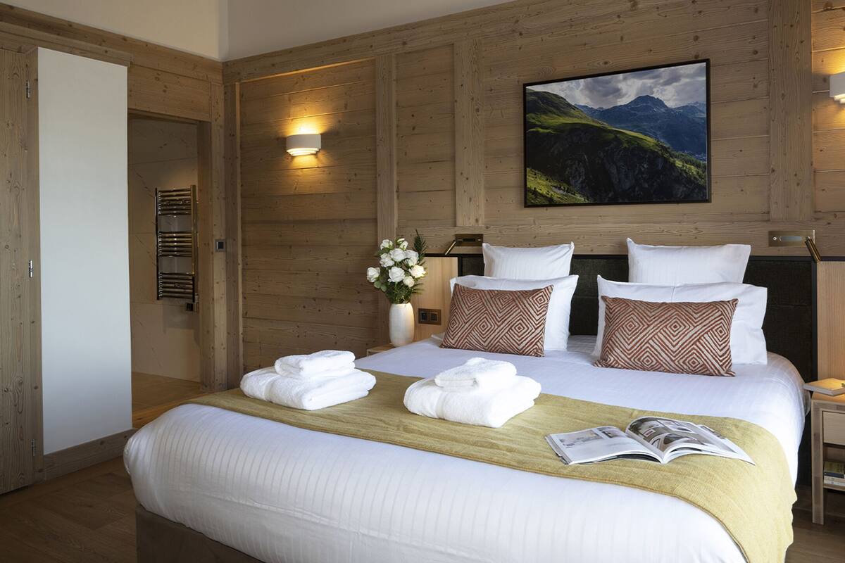 Ein Hotelzimmer mit holzgetäfelten Wänden, einem großen Bett mit weißer Bettwäsche, zwei Kissen, einem Nachttisch mit Lampe und einem Gemälde einer Berglandschaft.