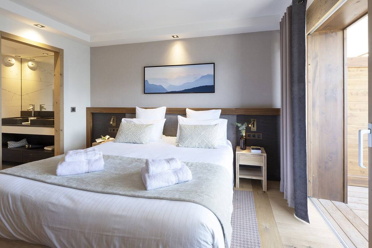 Ein modernes Hotelzimmer mit einem großen Bett, einem hölzernen Kopfteil, einem Nachttisch mit Lampe und einem großen Fenster mit Blick ins Freie.