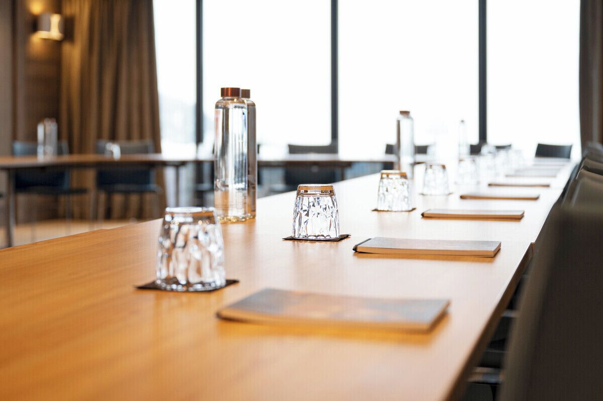 A conference room with a long table, clear glass tumblers, and a high ceiling with windows.