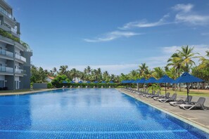 A swimming pool with lounge chairs and umbrellas, surrounded by palm trees and a building with balconies.