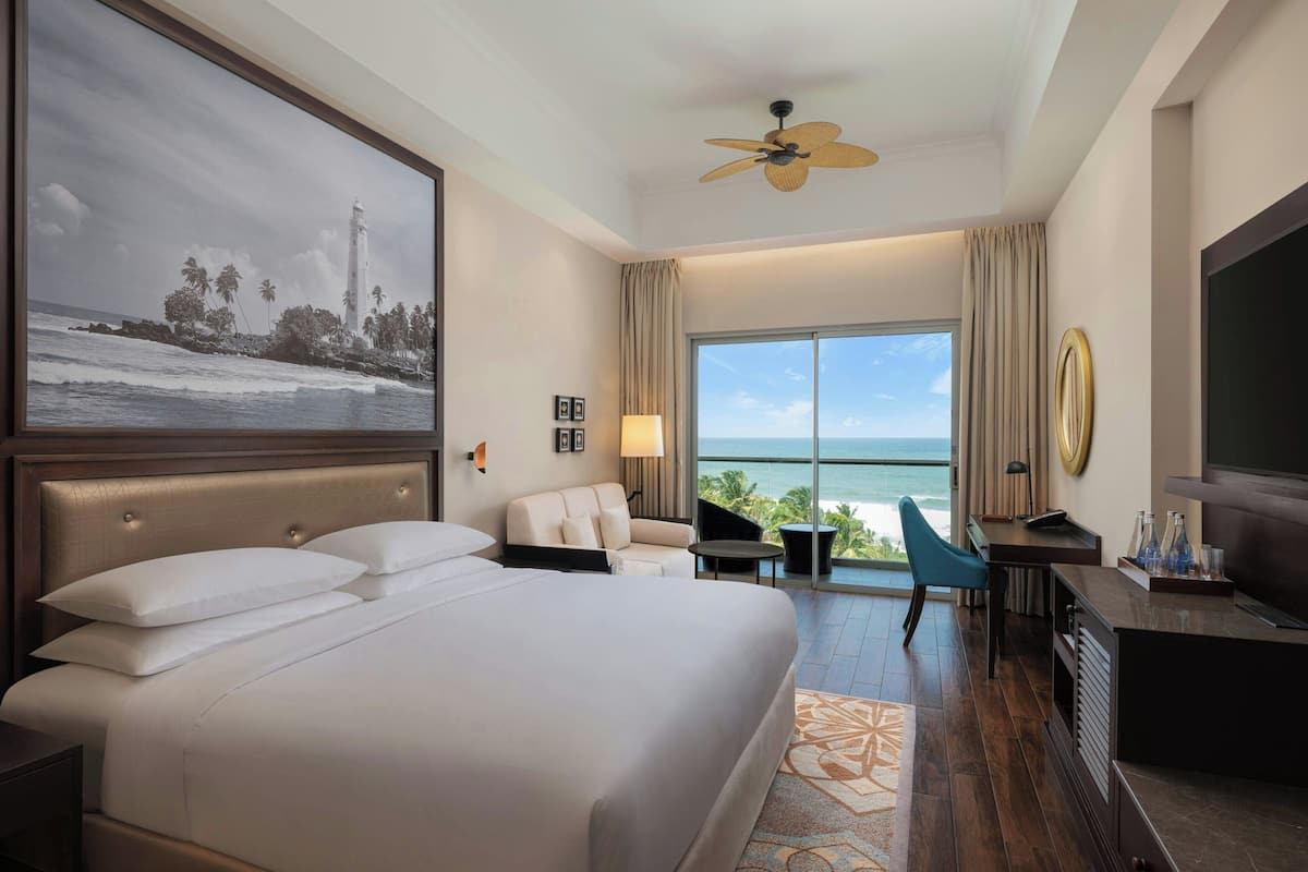 Ein Hotelzimmer mit einem großen Bett, einem Schreibtisch, einem Sessel, einem Fernseher, einem Deckenventilator und einem großen Fenster mit Blick aufs Meer.