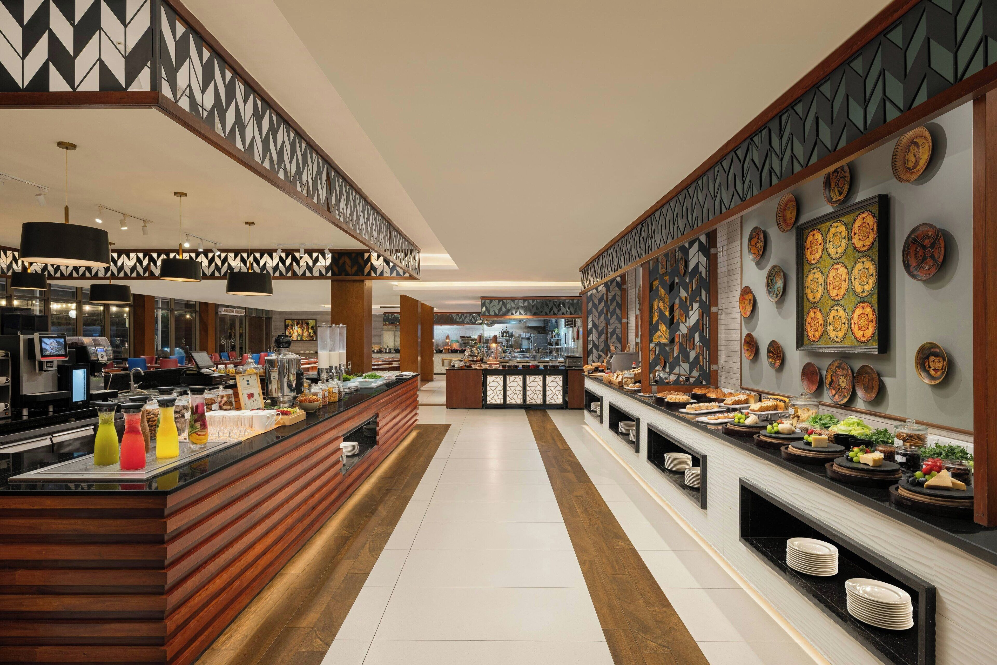 A modern hotel lobby with a long counter displaying food and beverages, a reception area, and a view into the kitchen.