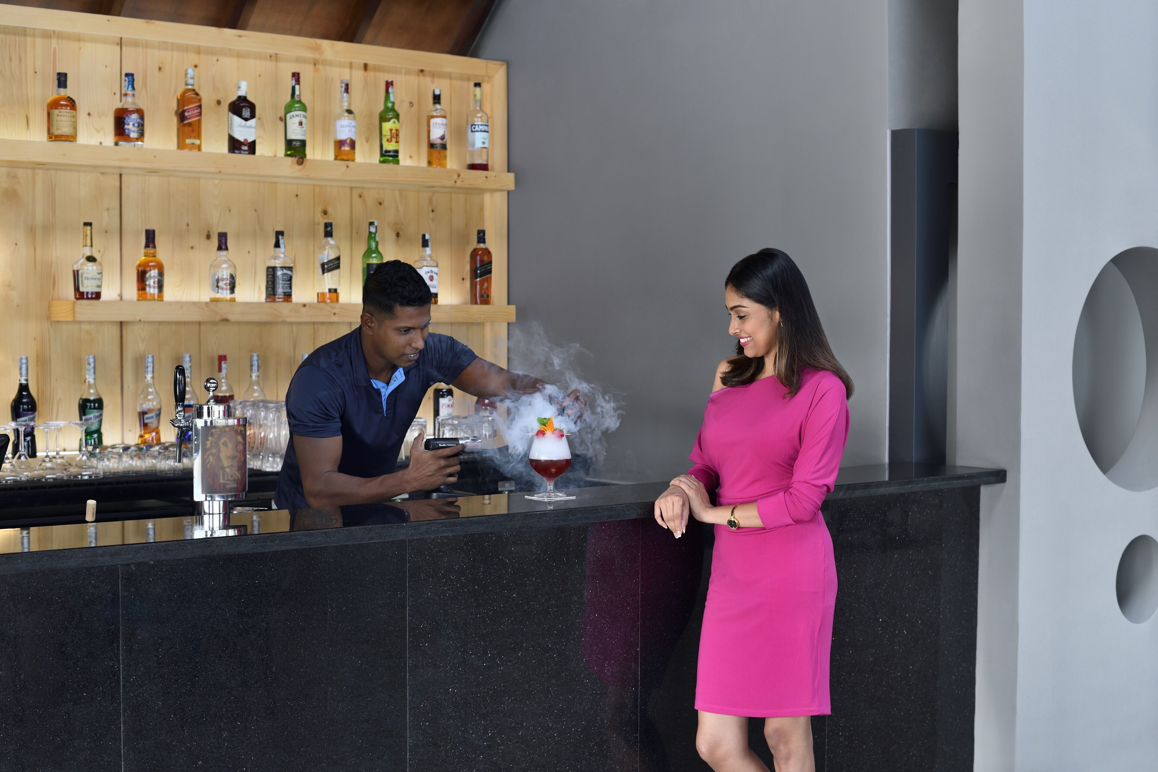 Zona de bar amb un barman preparant una beguda, una dona amb un vestit rosa i estanteries plenes de diferents ampolles de licors.