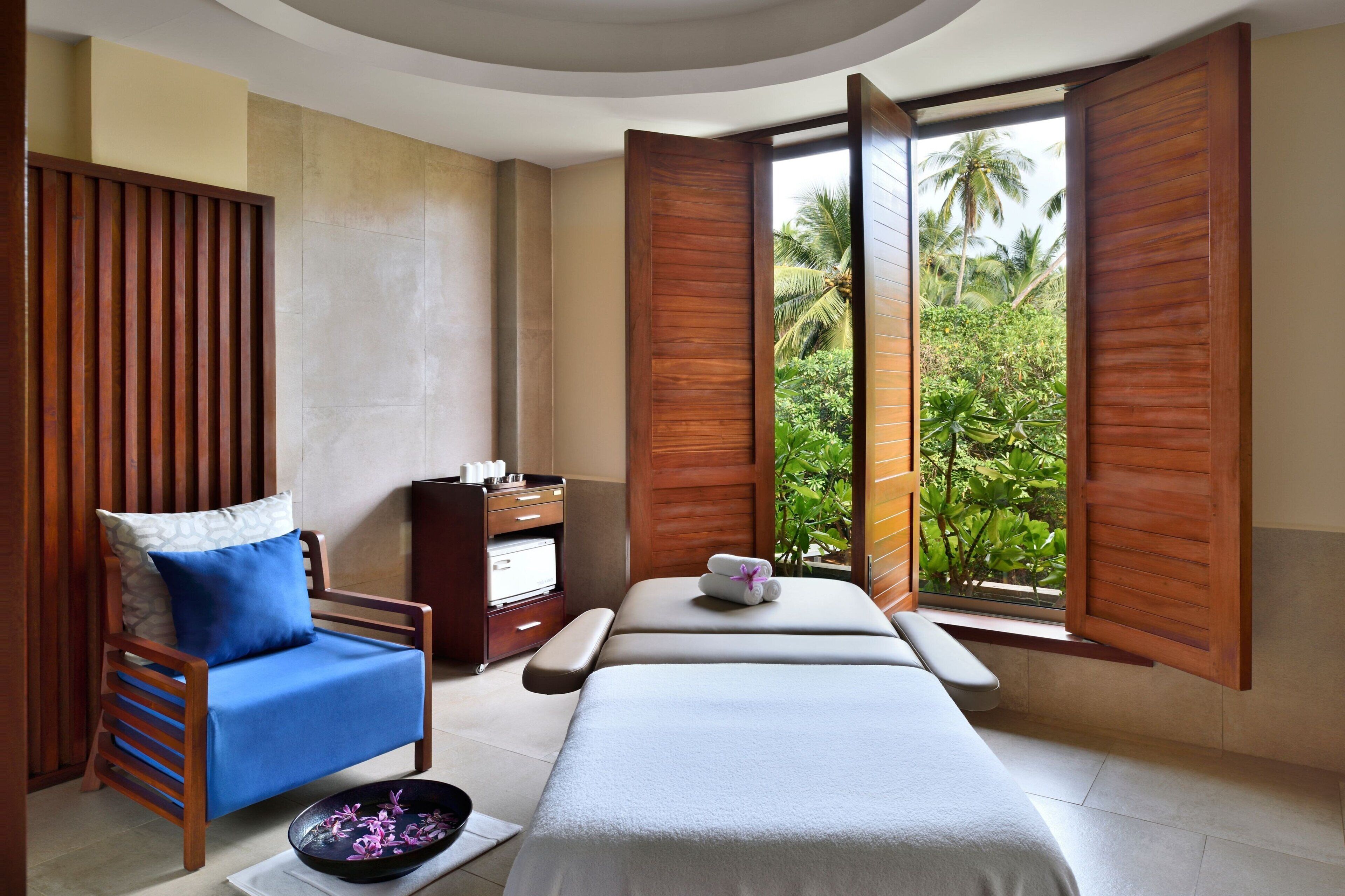 A spa room with a massage table, a wooden chair with a blue cushion, and a view of tropical greenery through open wooden shutters.