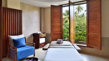 A spa room with a massage table, a wooden chair with a blue cushion, and a view of tropical greenery through open wooden shutters.