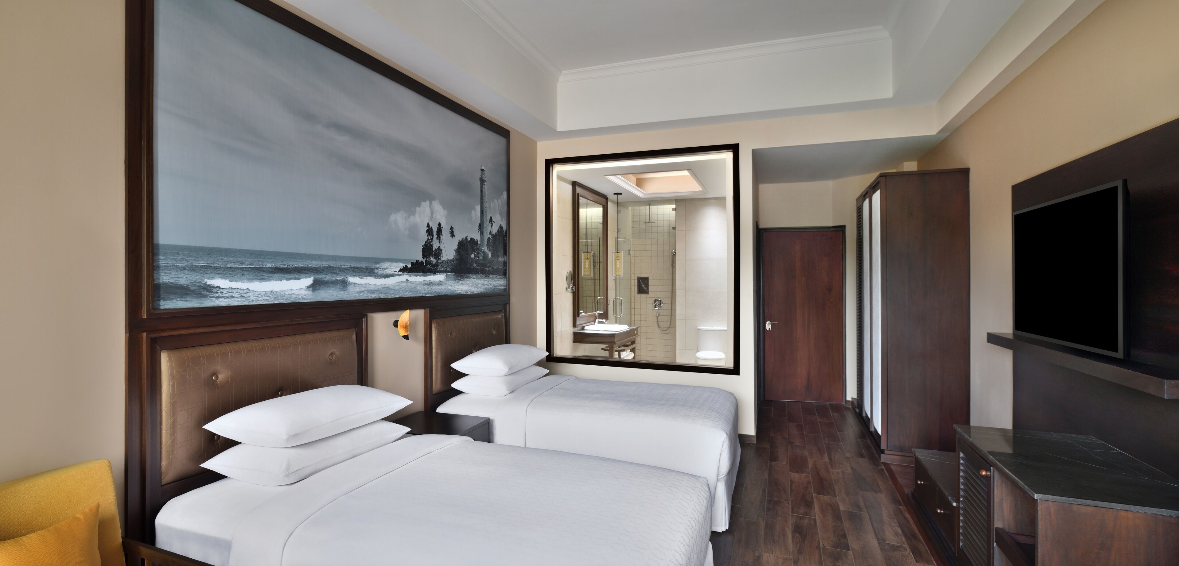 A hotel room with two beds, a large painting of a lighthouse, a TV, and a mirror reflecting the bathroom.