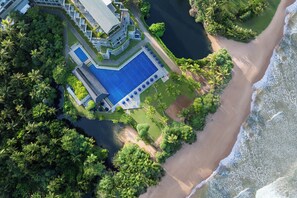 An aerial view of a resort with a large swimming pool, beachfront, and lush greenery.