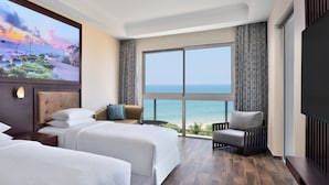 Ein Hotelzimmer mit zwei Betten, einem großen Fenster mit Meerblick, einem Sessel und einem Flachbildfernseher.