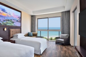 Habitación de hotel con dos camas, un ventanal con vistas al océano, una silla y un televisor de pantalla plana.
