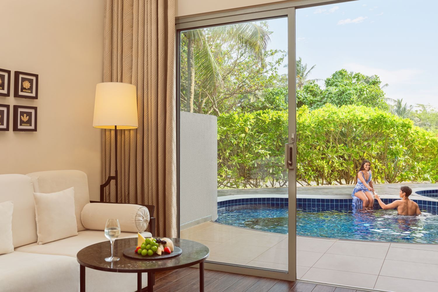A room with a sofa, a table with fruit and a glass, and a view of a swimming pool with people.