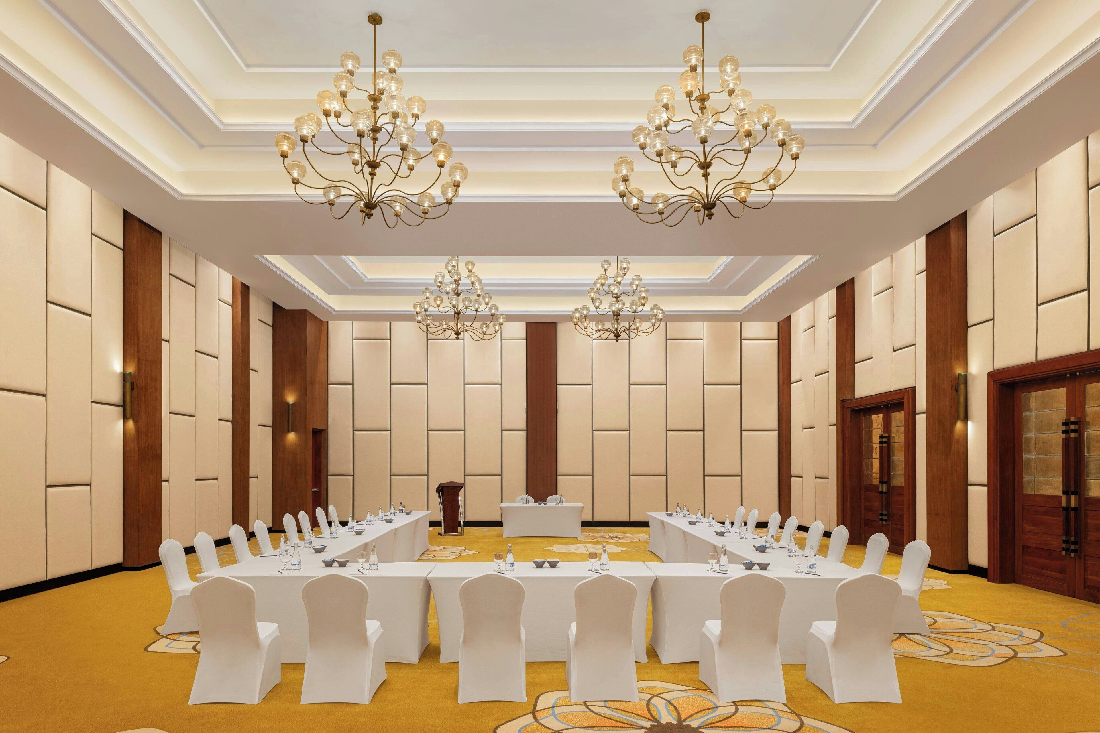 A conference room with a long table, white chairs, chandeliers, and a podium.