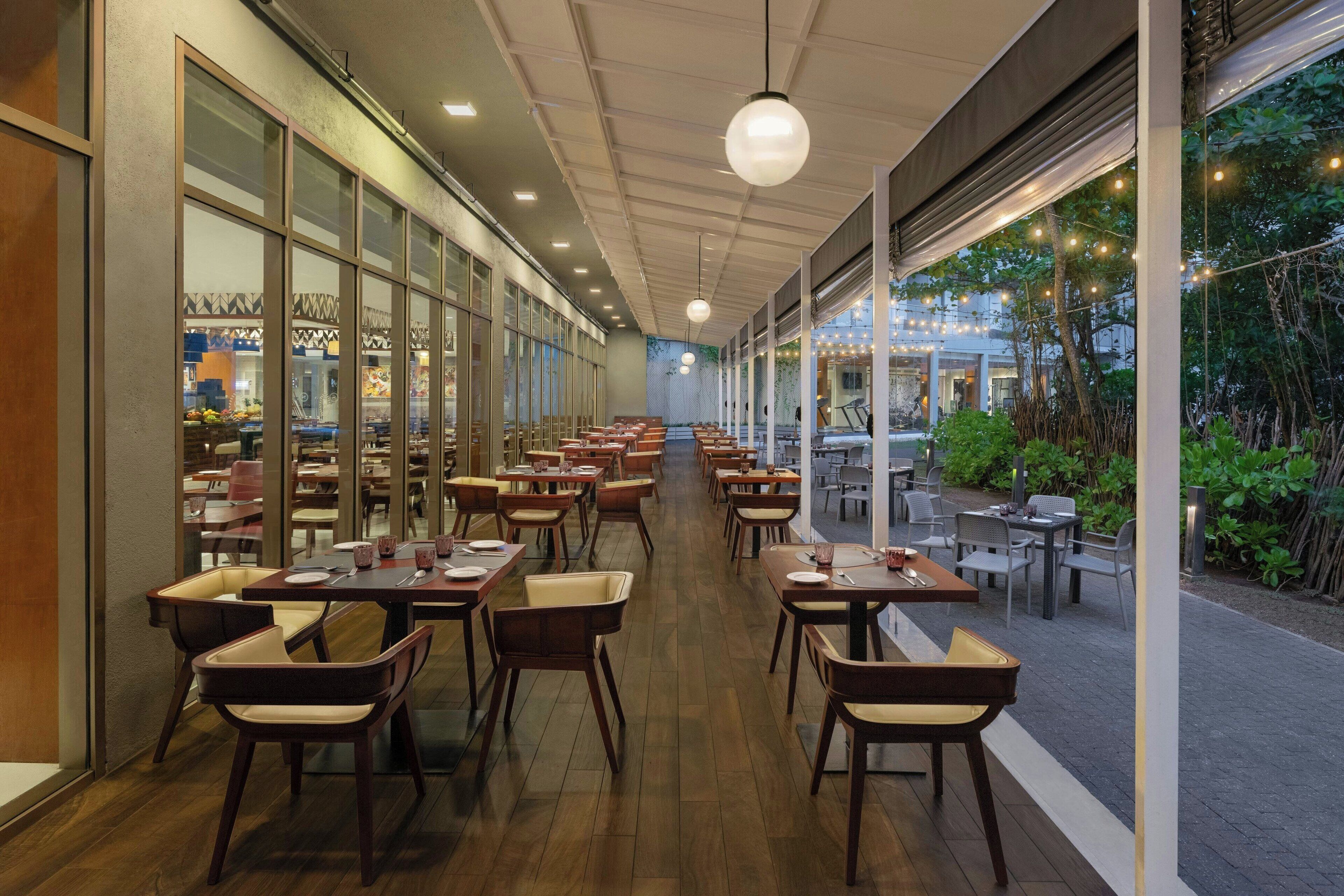 A spacious indoor dining area with multiple tables and chairs, large windows, and a view of the outdoor garden.