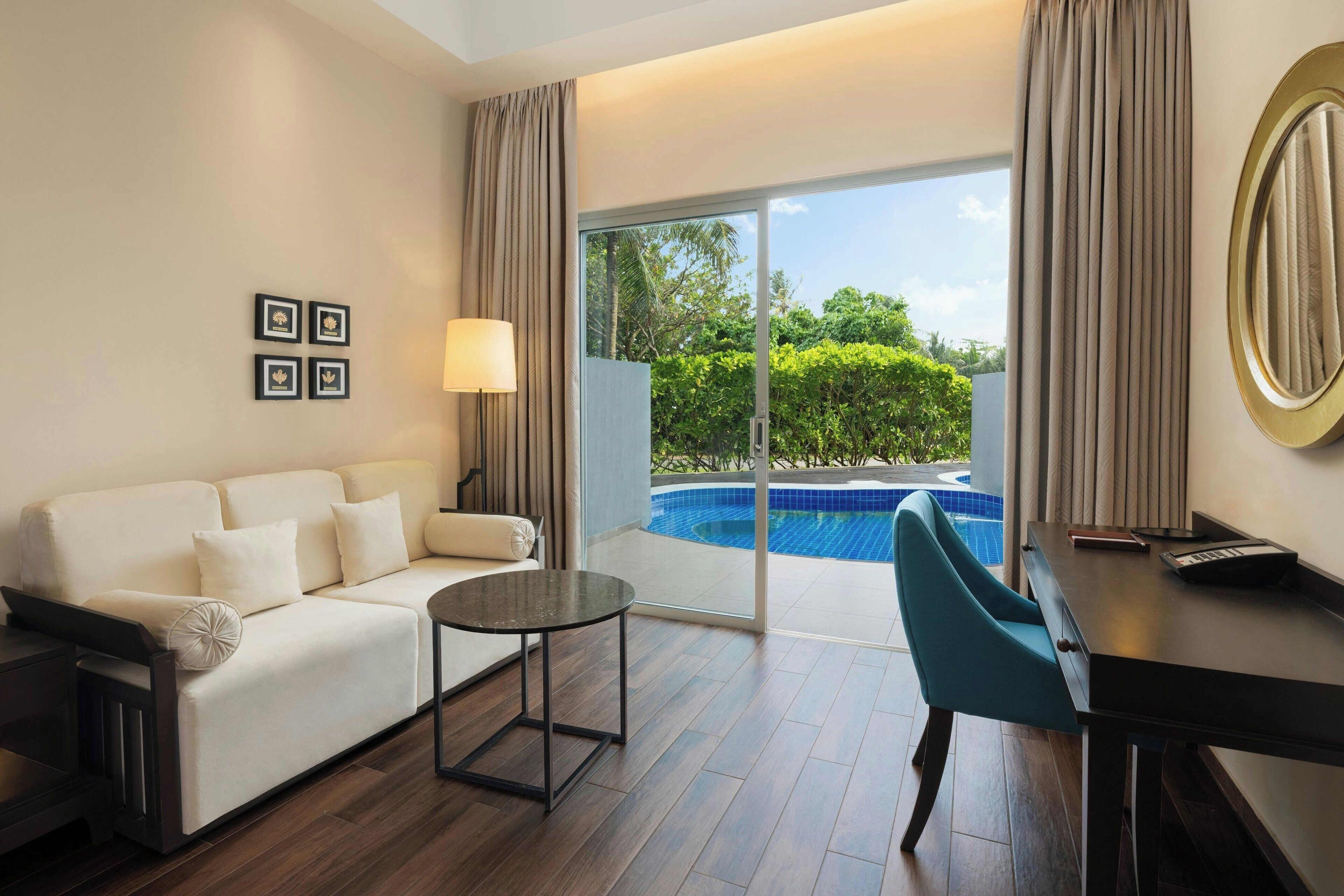 Habitación de hotel con sofá, una mesita, escritorio, ordenador portátil, teclado, lámpara y vistas a una piscina.