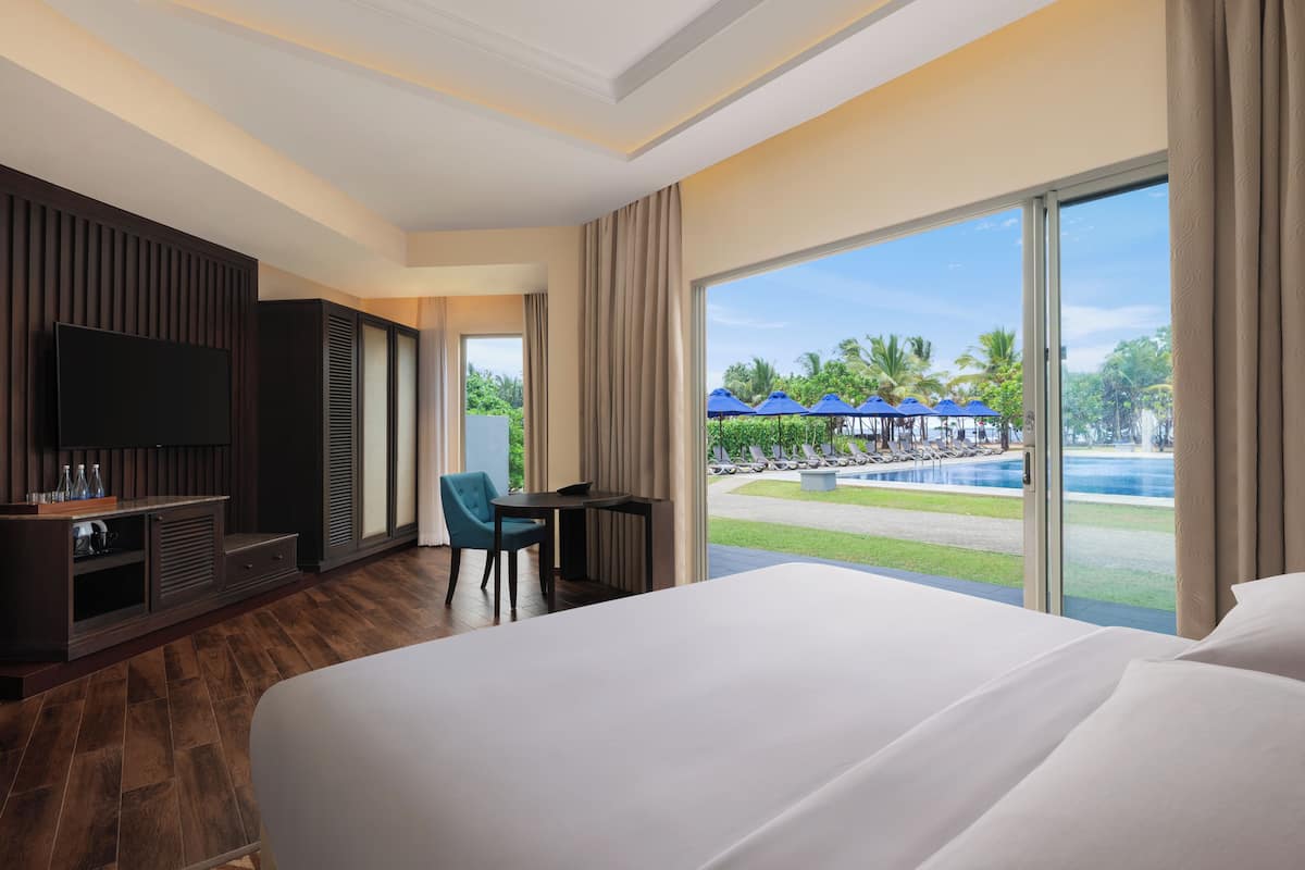 Ein Hotelzimmer mit einem großen Bett, einem Fernseher, einem Schreibtisch und Blick auf einen Pool und Palmen.