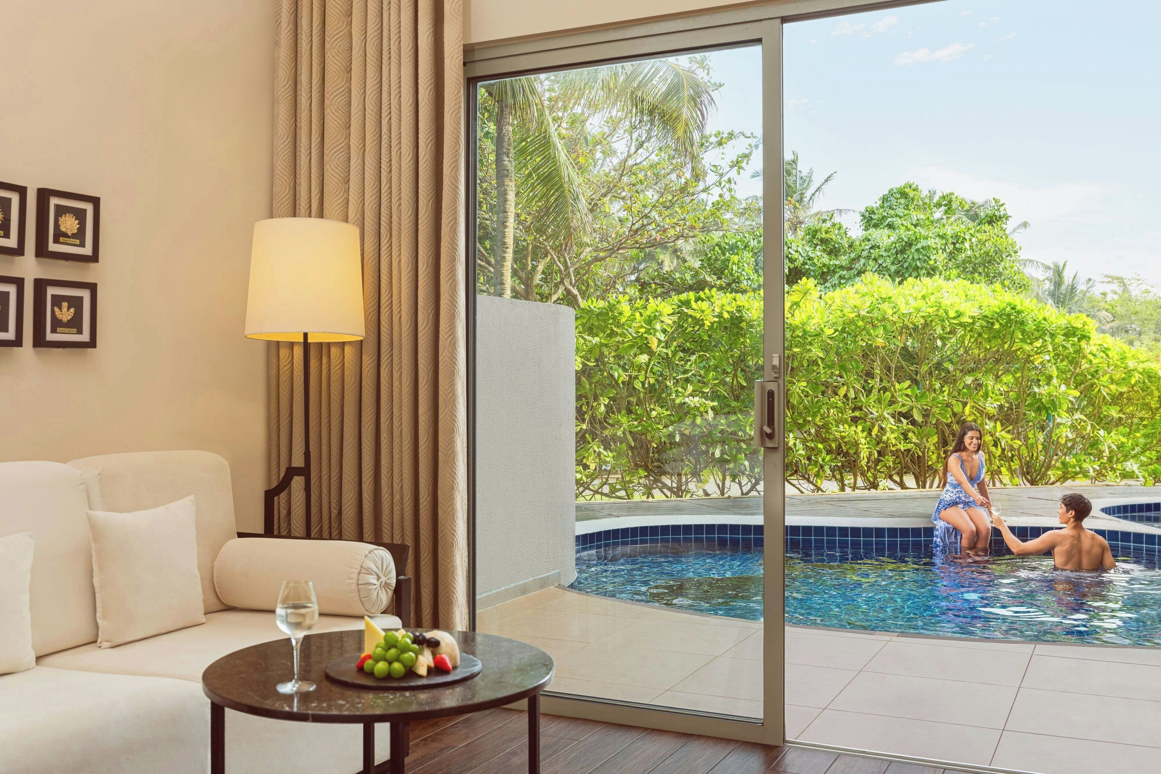 Une chambre d’hôtel moderne avec un canapé, une table avec des fruits et un verre de vin, et une vue sur une piscine où se trouvent deux personnes.