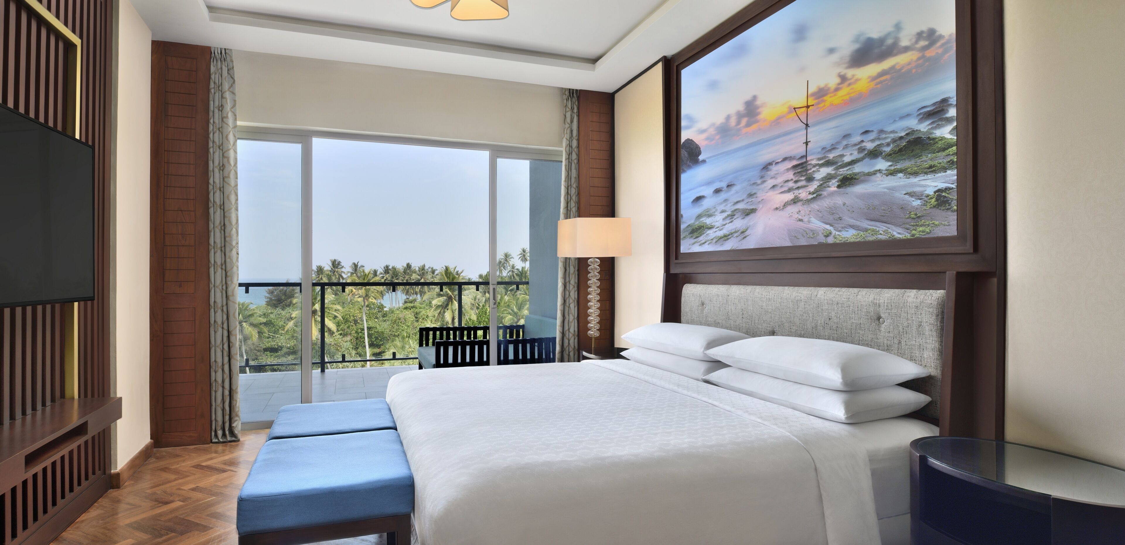 A hotel room with a large bed, a TV, a balcony with a view of palm trees, and a painting of a beach scene.