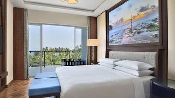 A hotel room with a large bed, a TV, a balcony with a view of palm trees, and a painting of a beach scene.