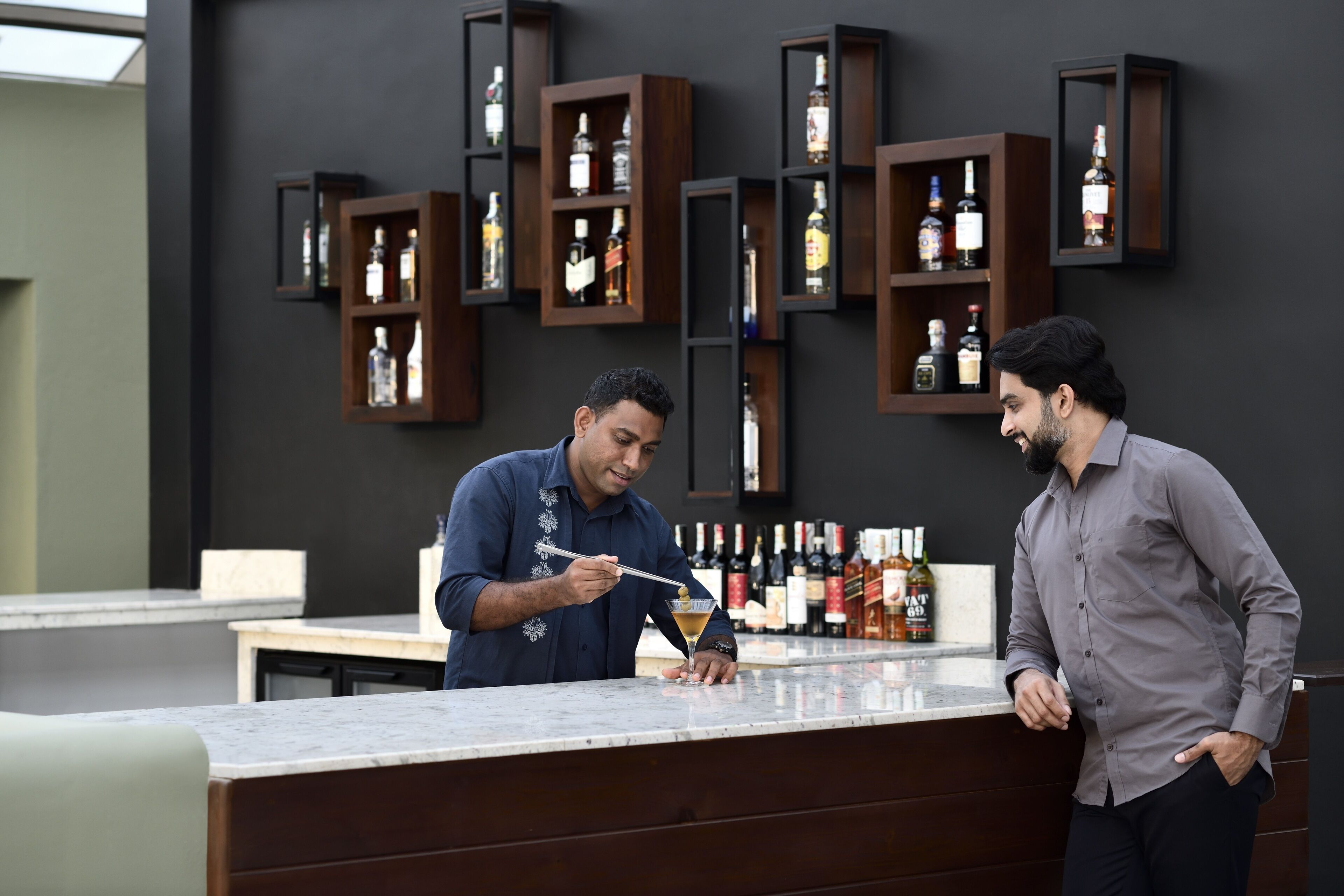 Dos hombres en un bar de diseño moderno, con estantes de madera oscura que exhiben botellas de licor.