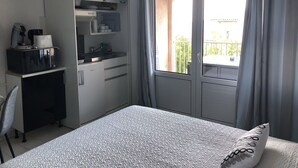 Een kleine hotelkamer met een bed, een kitchenette en een balkon met gordijnen.