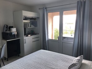 A compact hotel room with a bed, a kitchenette, and a balcony with curtains.