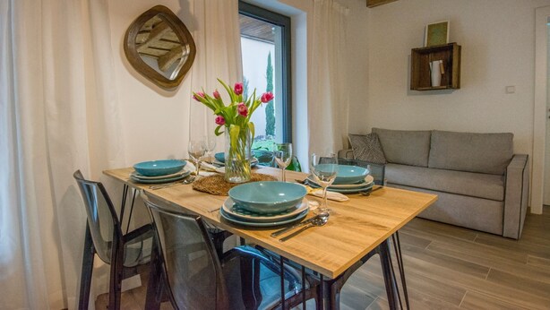 A dining area with a wooden table set for four, surrounded by chairs. A vase with flowers is on the table, and there is a window with curtains.
