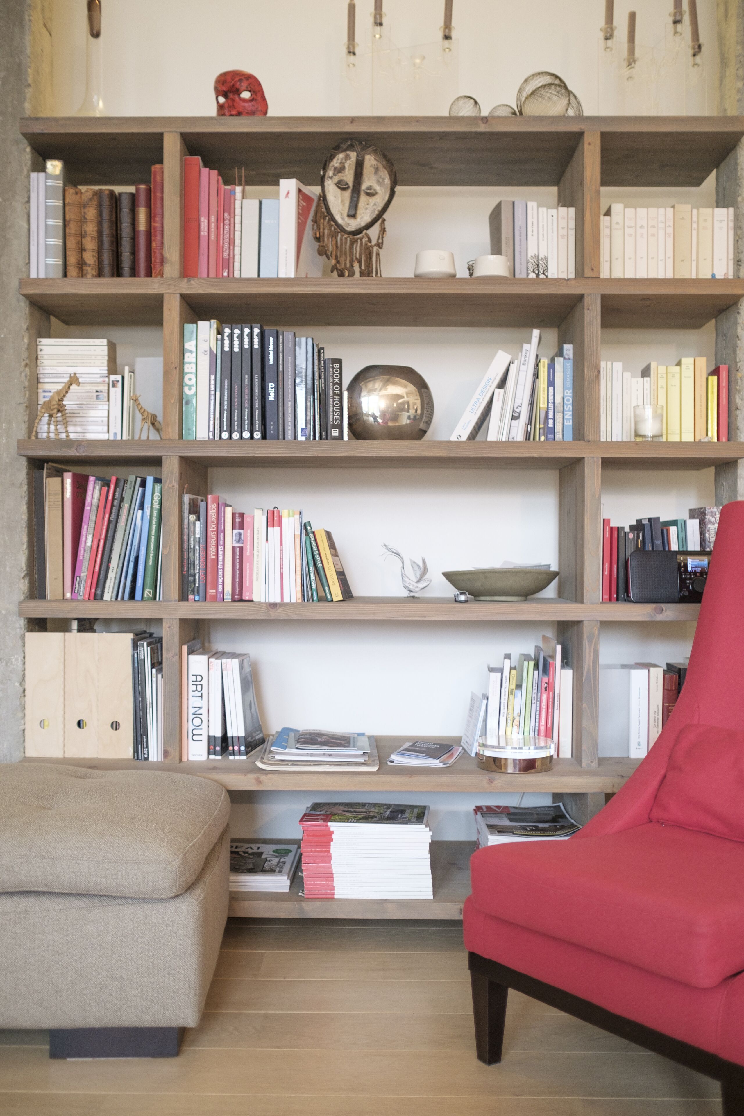 Une bibliothèque remplie de livres et d’objets décoratifs, un fauteuil rouge et une petite table sur laquelle se trouvent un bol et une horloge.