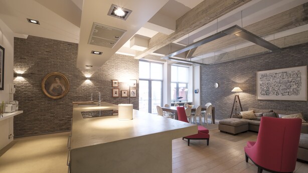 Una cucina moderna con isola centrale, una zona pranzo con una sedia rossa e un soggiorno con una grande finestra.