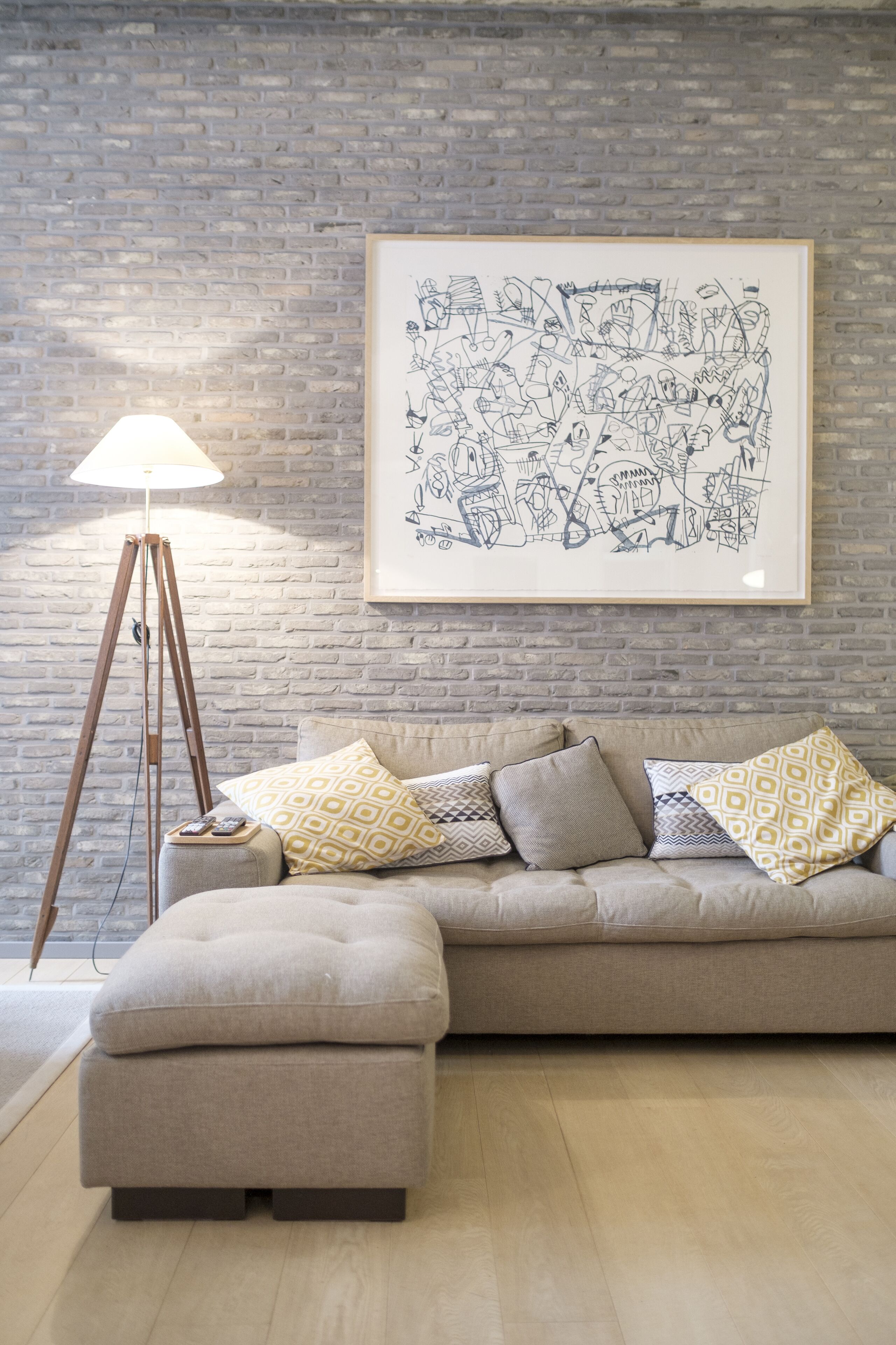 A modern living room with a brick wall, a floor lamp, a sofa with patterned cushions, and a framed abstract artwork.