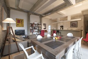 A spacious living area with a large wooden dining table, white chairs, a sofa, and a bookshelf.