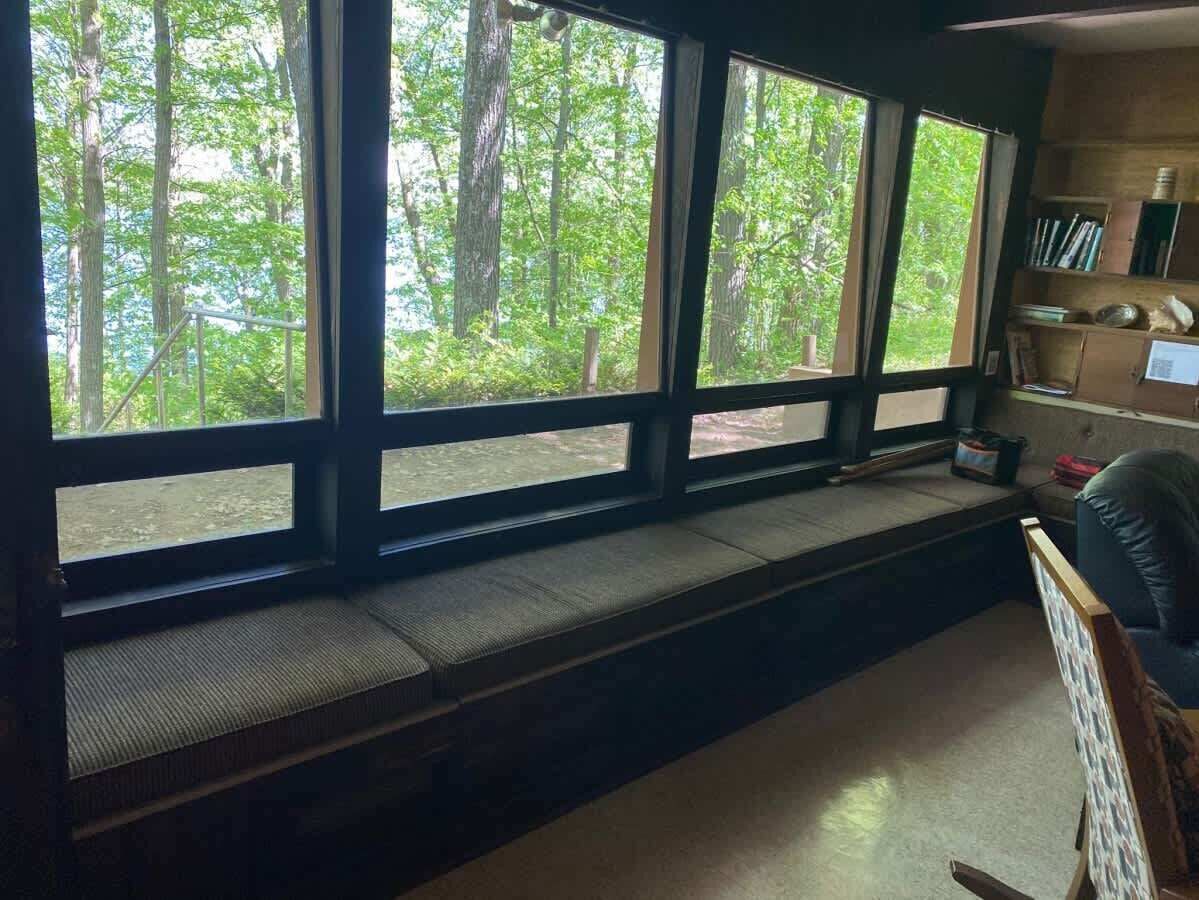 A room with a large window overlooking a forest, a built-in bench, and a bookshelf.