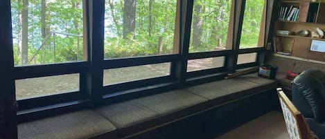 A room with a large window overlooking a forest, a built-in bench, and a bookshelf.
