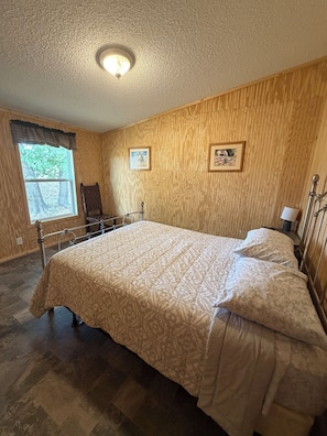 Un dormitorio con una cama grande, paredes de paneles de madera, un ventanal con vistas a árboles y dos cuadros enmarcados.