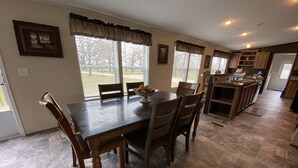 A dining area with a dark wooden table and chairs, a bowl of fruit, and a view of the outdoors through large windows. - Texas Ranch House on 10 acres near Lake Ray Roberts! (Pilot Point)