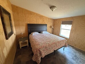 Une chambre avec un mur lambrissé en bois, un lit recouvert d’une couvre-lit à motifs floraux, une table de chevet, une lampe et une fenêtre avec des stores.