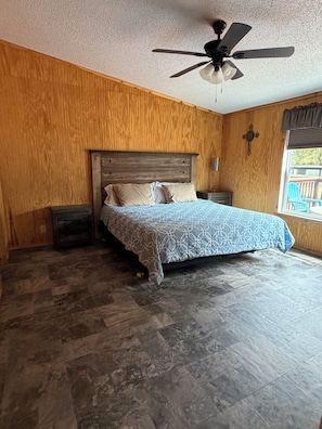 A bedroom with a bed, wooden walls, a ceiling fan, and a view of the outdoors.