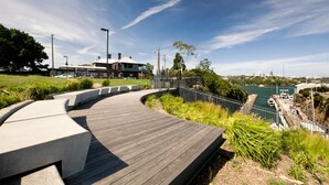 A curved wooden boardwalk with a concrete edge, surrounded by greenery and overlooking a body of water with boats.